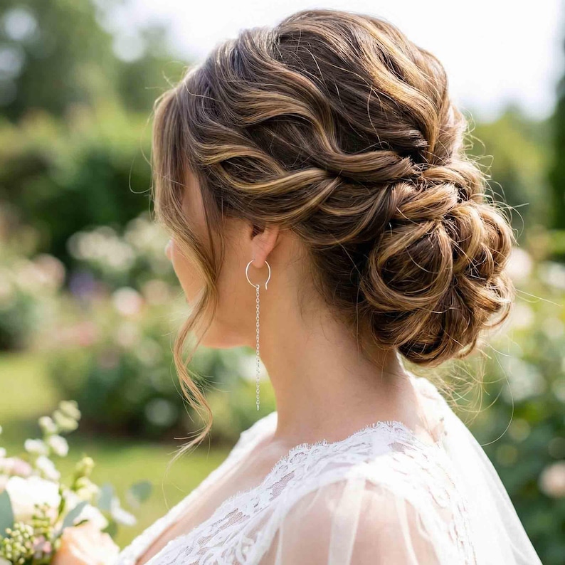 May include: A woman with a sophisticated hairstyle, wearing silver chain earrings. The hair is styled in a low bun with loose waves. The woman is wearing a white lace dress, with a blurred green background.