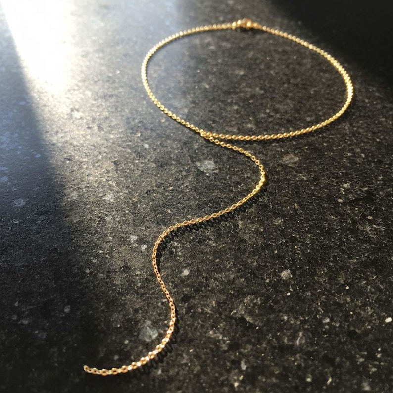 May include: A delicate gold-colored necklace with a circular design and a dangling chain. The necklace is made of small, connected links and rests on a dark, textured surface. The chain hangs down, creating a simple, elegant look.