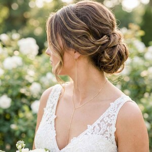 May include: A woman with her hair styled in an updo, wearing a white lace dress and a gold necklace. The necklace has a long, delicate chain that hangs down the front of the dress. The background is blurred, with white flowers and greenery.