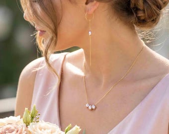 May include: A gold chain necklace featuring three small, light pink teardrop-shaped pearls at the center, paired with a matching gold hoop earring that has a delicate gold chain drop and a single small, light pink teardrop-shaped pearl accent.