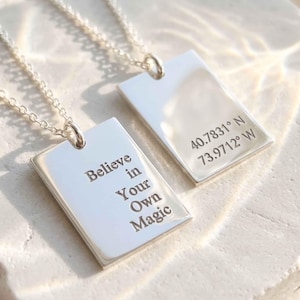 May include: Two silver-toned rectangular pendant necklaces with delicate chains. One pendant is engraved with the text "Believe in Your Own Magic" and the other pendant is engraved with the coordinates "40.7831° N 73.9712° W" in a clean font.