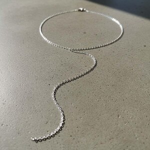 May include: A delicate silver Y-necklace with a simple, elegant design. The necklace features a fine chain that forms a circle around the neck, with a longer chain extending down. The jewelry is displayed on a gray surface.