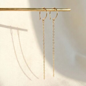 May include: A pair of delicate gold chain earrings. The earrings hang from a gold hook and feature a long, slender chain. The earrings are displayed on a gold bar against a soft, neutral background.