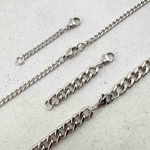 May include: Close-up of several silver-colored chain extenders with lobster claw clasps. The chains vary in thickness and length, with a polished, reflective surface. The extenders are designed for jewelry making and are displayed against a light gray background.