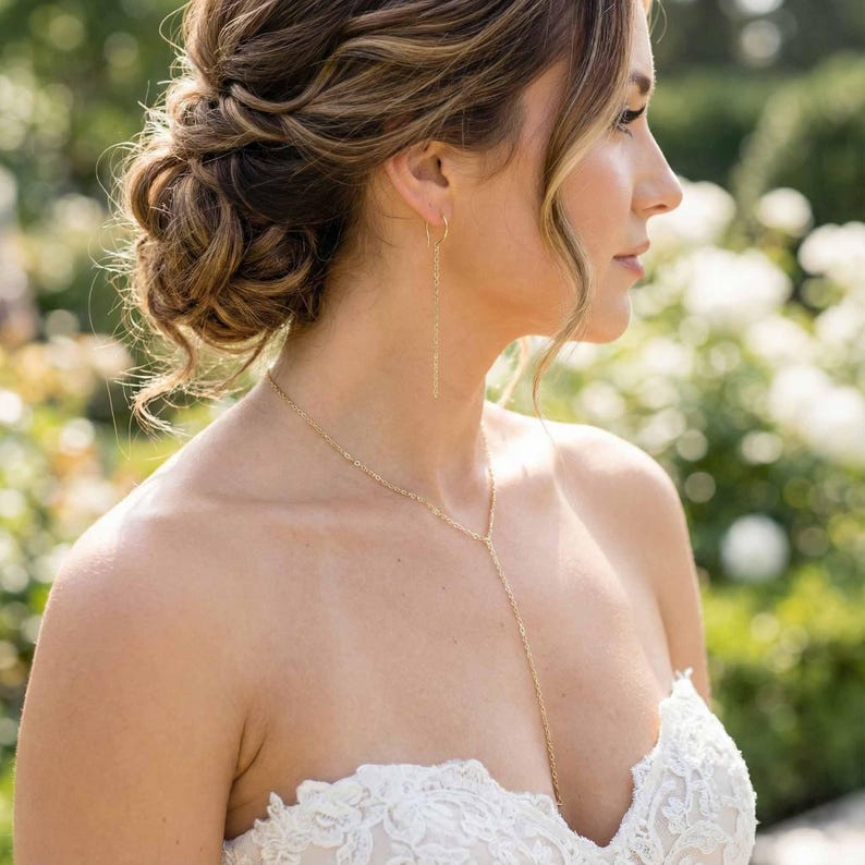 May include: A gold-colored necklace and matching earrings. The necklace has a delicate chain that drapes down the chest. The earrings are long and slender, hanging from the ear. The woman is wearing a white strapless dress with lace detailing.