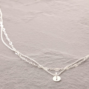 May include: A delicate silver necklace with two strands. The top strand features oval-shaped links, while the bottom strand has a chain with a small, round pendant. The pendant is engraved with the letter "k".