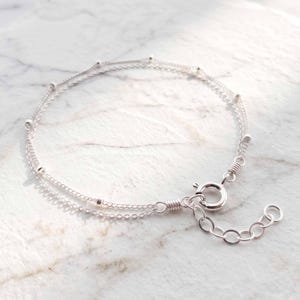 May include: A double-strand silver bracelet featuring delicate chain links interspersed with small, evenly spaced silver beads. The bracelet is secured with a round spring ring clasp and includes an adjustable extender chain with circular links.