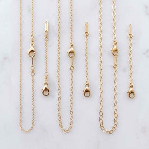 May include: Three gold chain necklaces arranged vertically, each paired with a matching gold chain extender. The necklaces feature delicate oval links and lobster claw clasps, while the extenders include small tags marked 14K GF.