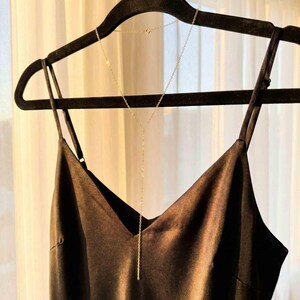 May include: A gold-colored Y-shaped necklace with a delicate chain hangs on a black hanger. The necklace is draped over a black satin camisole top. The background is a sheer white curtain.