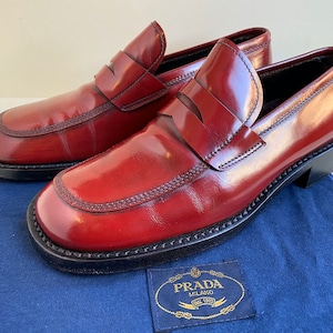 Puede incluir: Un par de mocasines de cuero burdeos con suela de cuero negro y un logotipo Prada en oro en relieve en la suela.