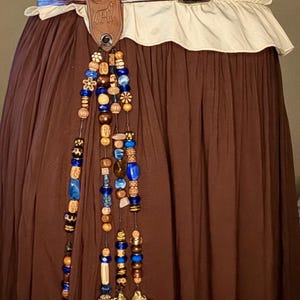 May include: A brown leather belt featuring an embossed wolf and moon design, worn over a cream-colored ruffled top and a brown skirt. Attached to the belt is a decorative accessory with multiple strands of blue, brown, and tan beads and a compass.