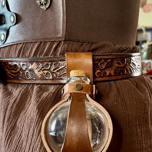 May include: A brown leather belt with embossed floral and dragon designs, holding a clear glass flask with a cork stopper. The belt is part of a brown outfit, with a decorative compass emblem.