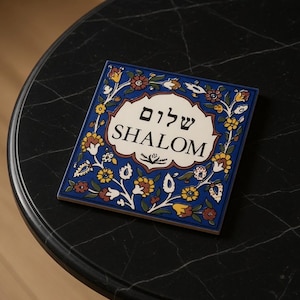 May include: Square ceramic tile with a blue floral border and the word "SHALOM" in English and Hebrew. The tile features a white center with the text and is decorated with yellow, red, and brown flowers.