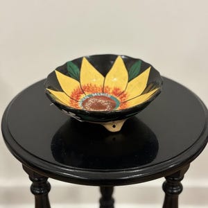 Peut inclure: Un bol en céramique peint à la main avec un motif de tournesol. Le bol présente des pétales jaunes, un centre marron et le mot "MEXICO" au centre. Le bol est posé sur une table d'appoint ronde noire.