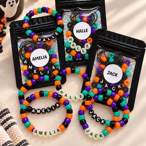 Peut inclure: Trois sachets noirs refermables avec des fenêtres holographiques, contenant chacun des perles en plastique colorées orange, violettes, noires et bleu sarcelle. Chaque sachet porte une étiquette circulaire blanche avec les noms AMELIA, HALLE ou JACK.