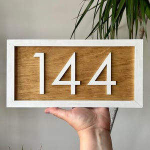 May include: Wooden house number sign displaying the number 144 in white, set against a light brown wood grain background. The numbers are raised and framed by a white border. A decorative piece for home decor.