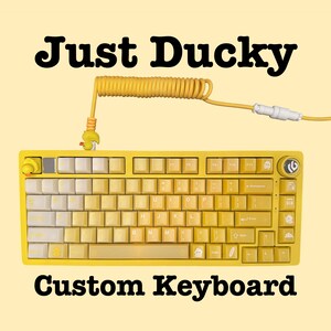 May include: Yellow custom keyboard with white and yellow keys. The keyboard has a coiled yellow cable and a duck-shaped charm. The text "Just Ducky" and "Custom Keyboard" are displayed.
