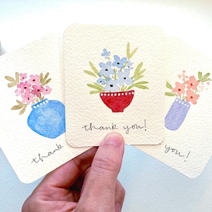 May include: Three off-white rectangular cards featuring watercolor illustrations of flowers in vases. The center card displays blue flowers in a red bowl with the text thank you! written below. The other cards show pink and orange floral arrangements.