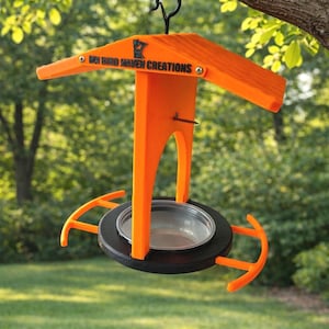 May include: An orange bird feeder with a black metal hook, hanging from a tree branch. The feeder has a roof, a central support, and a circular base with a clear bowl. The text "MY BIRD HAVEN CREATIONS" is printed on the feeder.
