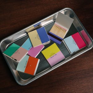 Peut inclure: Une collection de boîtes d'allumettes carrées aux combinaisons de couleurs variées, notamment bleu, jaune, rose, vert et orange, avec le texte "the smiths" sur les couvercles. Une boîte ouverte révèle des allumettes en bois à bouts roses.