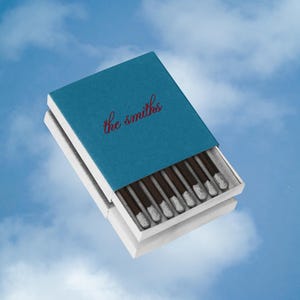 Peut inclure: Une boîte d'allumettes rectangulaire avec un étui coulissant bleu sarcelle portant l'inscription the smiths en écriture cursive rouge. La boîte est entrouverte, révélant une rangée d'allumettes brun foncé aux bouts gris clair.