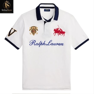 Peut inclure: Polo blanc avec col et bord des manches bleu marine. Il présente le texte brodé "Ralph Lauren" en bleu, un écusson et un logo de joueur de polo rouge. La manche gauche porte un "V" bleu marine avec une bordure dorée.