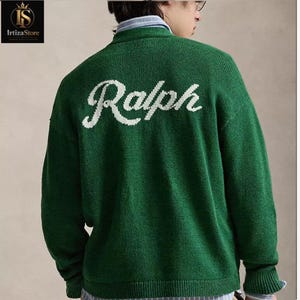 Peut inclure: Un pull vert en tricot avec le nom "Ralph" en écriture blanche sur le dos. Le pull a un col montant et des manches longues. Une chemise rayée est visible en dessous.