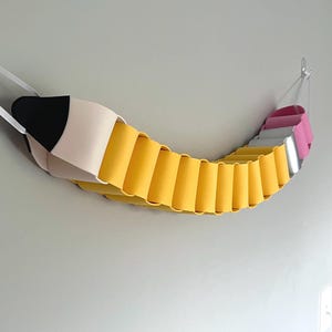 May include: A whimsical, oversized pencil-shaped wall decor piece. The pencil features a black tip, a beige body, and a yellow barrel. The eraser end is pink and silver. The piece is suspended from the wall with white ribbons.