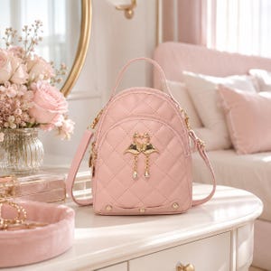 May include: A light pink quilted backpack featuring a top handle and two shoulder straps. The front is adorned with a gold-toned metal emblem of two kissing flamingos with dangling chain accents and small clear crystals at the base of the chains.