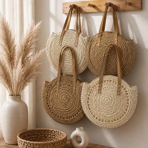 May include: Four round woven straw bags with tan leather handles hanging on a wooden wall rack. The bags feature circular patterns in shades of beige and light brown. Each bag has two straps attached with visible stitching at the top edge.