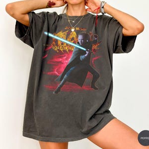 May include: Dark gray t-shirt featuring a graphic of a character with a blue lightsaber, set against a red and yellow background with the word "SHADOW". The shirt has a vintage, worn look.