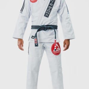 May include: A white Brazilian Jiu-Jitsu gi featuring the Gracie Barra logo in red and white on the chest and trouser leg. The jacket includes a black lapel patch with the text Gracie Barra, black sleeve stripes, and a dark blue belt tied at the waist.