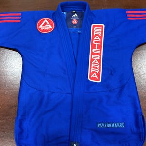 Puede incluir: Un gi de artes marciales azul con detalles de tres rayas rojas en los hombros, un parche circular de Gracie Barra en el pecho derecho, un logotipo vertical de Gracie Barra en la solapa izquierda y una etiqueta azul de Performance abajo.
