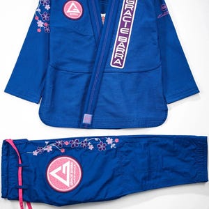 Puede incluir: Un conjunto de kimono de Jiu-Jitsu brasileño azul que incluye chaqueta y pantalón. La chaqueta presenta un parche con el logotipo de Gracie Barra, bordados de flores de cerezo rosas en el hombro y un parche de texto vertical Gracie Barra.