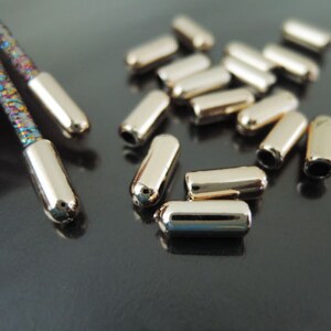 Puede incluir: Extremos de cordón de metal dorado y una sección de un cordón multicolor. Los extremos del cordón son cilíndricos con una parte superior redondeada y un agujero en la base. Varios extremos de cordón sueltos están esparcidos sobre una superficie oscura.