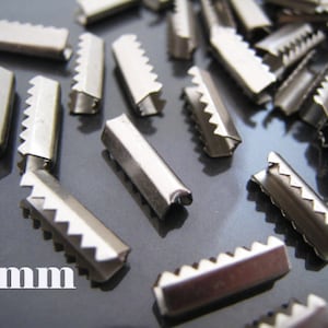 May include: A close-up of a pile of silver metal slider buckles, each measuring 2mm in width. The buckles have a serrated edge and are designed for attaching straps or cords.