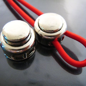 Puede incluir: Dos cierres de botón de metal plateado con un cordón elástico rojo enhebrado. Los cierres tienen una superficie lisa y redondeada y un borde ligeramente elevado.