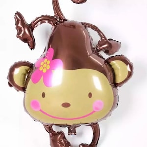 May include: A brown and gold foil balloon shaped like a monkey with a pink flower on its head. The monkey has a smiling face with pink cheeks and brown arms and legs.