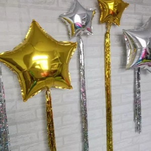 Può includere: Quattro palloncini a forma di stella in lamina con nastri dorati e argentati attaccati. I palloncini sono appesi a un muro.