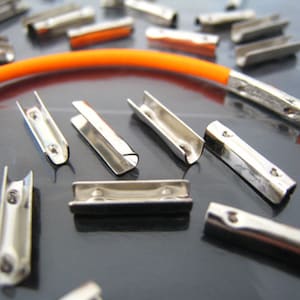K&ouml;nnte beinhalten: Nahaufnahme einer Gruppe von silbernen Metall-Kordelenden, von denen einige auf eine orangefarbene elastische Kordel gef&auml;delt sind. Die Kordelenden sind so konzipiert, dass sie zum Befestigen von Kordeln oder B&auml;ndern verwendet werden k&ouml;nnen.