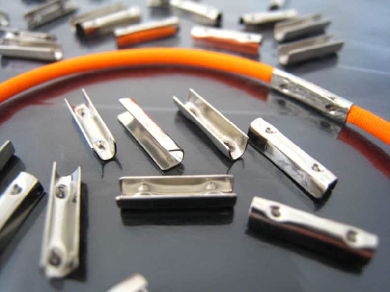 K&ouml;nnte beinhalten: Nahaufnahme einer Gruppe von silbernen Metall-Kordelenden, von denen einige auf eine orangefarbene elastische Kordel gef&auml;delt sind. Die Kordelenden sind so konzipiert, dass sie zum Befestigen von Kordeln oder B&auml;ndern verwendet werden k&ouml;nnen.