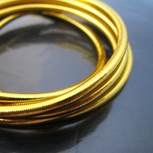 Puede incluir: Primer plano de un conjunto de cuatro pulseras de brazalete doradas. Las pulseras están apiladas una encima de la otra y tienen un acabado metálico brillante.