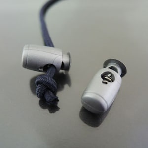 Puede incluir: Dos cierres de cordón de plástico plateado con un cordón negro enhebrado en uno de ellos. Los cierres de cordón se utilizan para ajustar la longitud de un cordón o una correa.