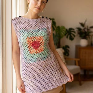 May include: A sleeveless, crochet top in a light purple hue, featuring a central square design with a rainbow gradient of colors. The top has a scoop neckline with three black buttons on the shoulder. The crochet pattern is open and airy.
