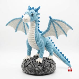 Puede incluir: Un juguete de dragón tejido a ganchillo en azul claro y blanco, de pie sobre una base de roca gris texturizada. El dragón tiene alas grandes con paneles interiores blancos, una cola larga con púas azules y vientre blanco. Icono PDF rojo.