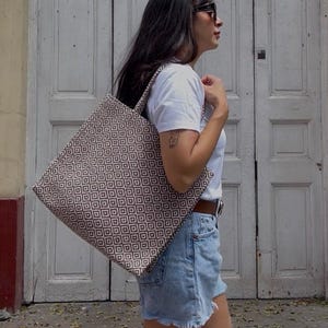 May include: A tote bag with a geometric pattern in brown and cream is carried over the shoulder. The bag is held by a person wearing a white t-shirt and denim shorts. The background features a white door and a yellow wall.
