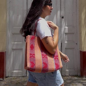 May include: A colorful tote bag with a geometric pattern in shades of orange, red, and pink. The bag is carried over the shoulder. The background features a white door and a yellow wall.