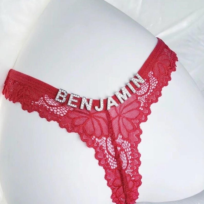K&ouml;nnte beinhalten: Roter Spitzen-String-Tanga mit dem Namen "BENJAMIN" in silbernen Strassbuchstaben &uuml;ber dem Bund. Die Unterw&auml;sche hat ein florales Spitzendesign und einen gewellten Rand.