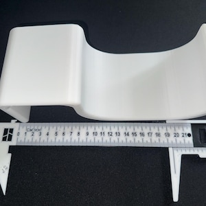 Op de afbeelding: Een witte, gebogen, U-vormige acryl displaystandaard geplaatst boven een witte plastic schuifmaat. De schuifmaat toont aan dat het object een breedte heeft van ongeveer 21 centimeter. De standaard heeft een plat bovengedeelte en een holle basis.