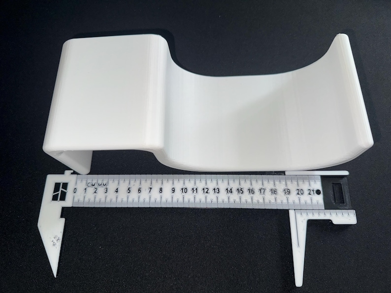 Op de afbeelding: Een witte, gebogen, U-vormige acryl displaystandaard geplaatst boven een witte plastic schuifmaat. De schuifmaat toont aan dat het object een breedte heeft van ongeveer 21 centimeter. De standaard heeft een plat bovengedeelte en een holle basis.